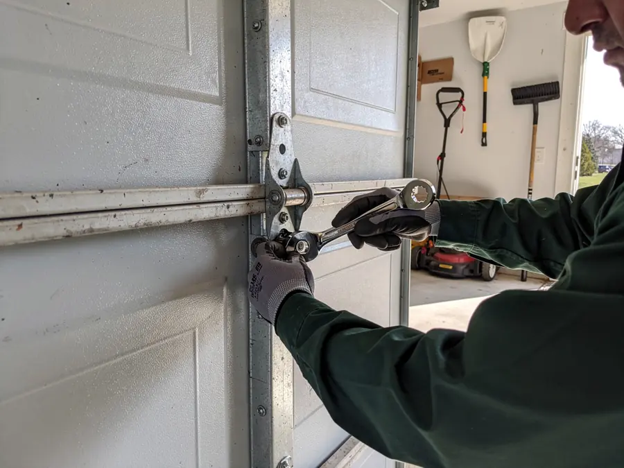 A technician in gloves uses a wrench to adjust a garage door hinge.